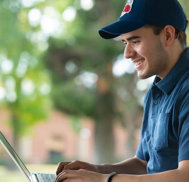 A student wearing an Ole Miss cap sits outside and looks at a laptop