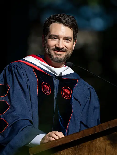 Bradford Cobb in graduation regalia smiling and standing behind a podium