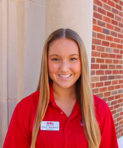 A School of Applied Sciences Ambassador smiles while standing outdoors beside a stone column on the University of Mississippi campus. She is wearing a red University of Mississippi School of Applied Sciences polo shirt and a name tag that reads "Alison Blackburn." A brick building and campus walkway are visible in the background.