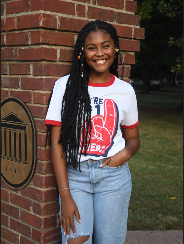 A young black woman smiles and leans against a brick column with one hand in the pocket of her jeans. She wears a ringer tee that reads "We're #1 Rebs" on the image of a foam finger.