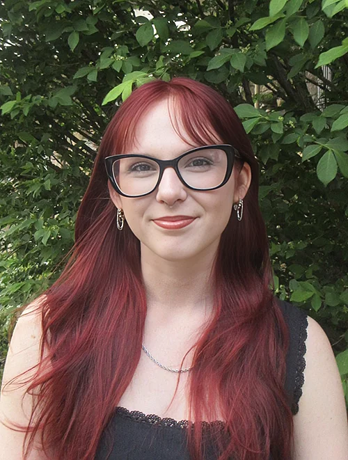 person with glasses and red hair standing outdoors smiling at camera
