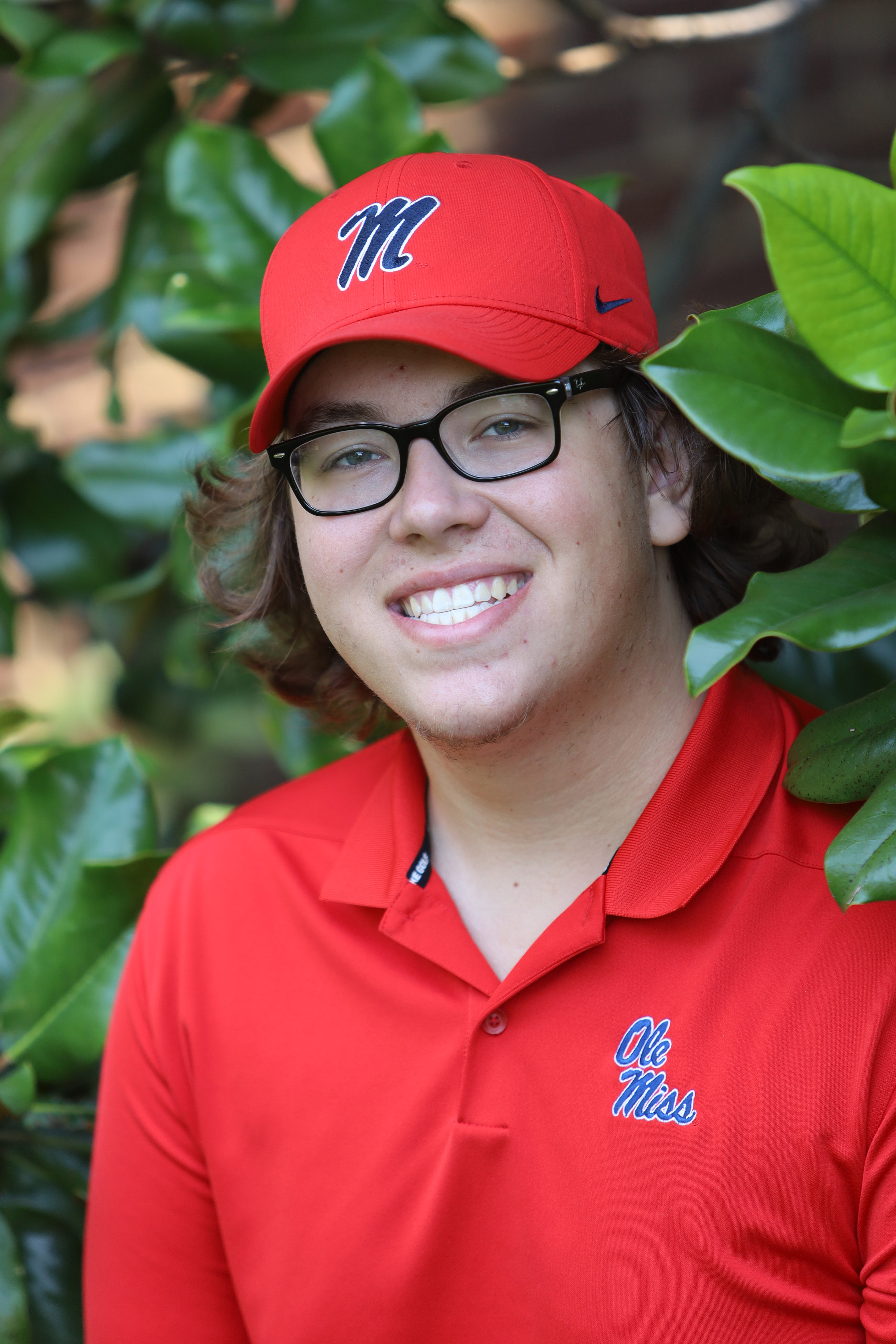 A young white man with glasses wearing red Ole Miss shirt and hat stands between Magnolia trees in bloom.