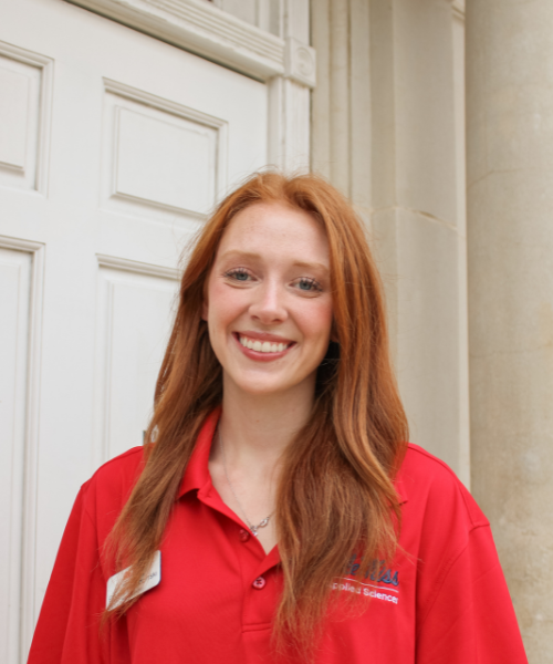A smiling School of Applied Sciences Ambassador stands outdoors in front of a white campus building entrance. She is wearing a red University of Mississippi School of Applied Sciences polo shirt and a necklace. Her long red hair falls over her shoulders, and stone columns frame the entrance in the background.