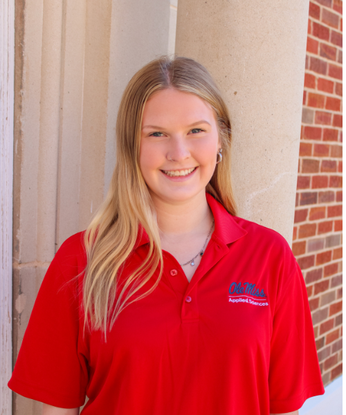 A School of Applied Sciences Ambassador smiles while standing outdoors beside a stone column on campus. She is wearing a red University of Mississippi School of Applied Sciences polo shirt with the embroidered Ole Miss logo and a necklace. A brick building and campus walkway are visible in the background.