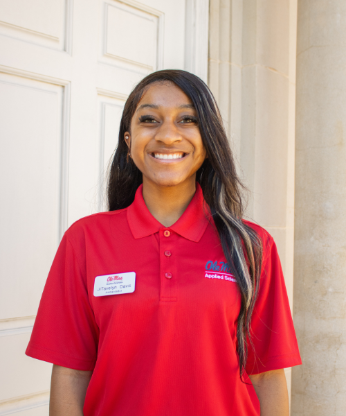 A smiling School of Applied Sciences Ambassador stands outdoors in front of a white campus building entrance. She is wearing a red University of Mississippi School of Applied Sciences polo shirt with an embroidered Ole Miss logo and a visible name tag that reads "JiTavelyn Davis." She faces the camera with long dark hair over one shoulder, and a stone column is visible beside the doorway in the background.