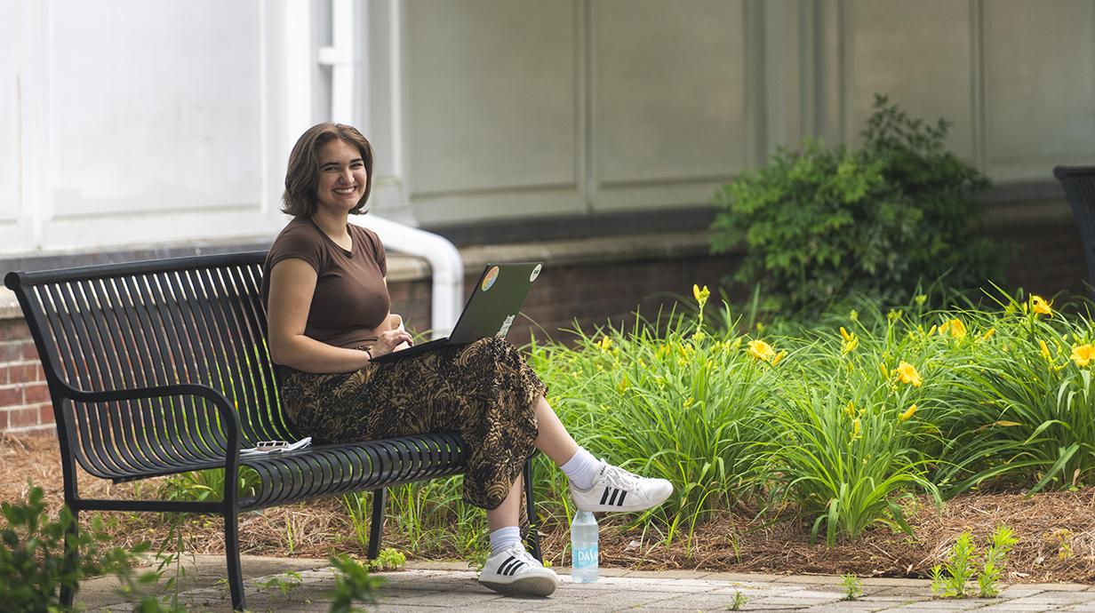 A student sits on an outdoor bench using a laptop beside a landscaped garden with yellow flowers, smiling toward the camera.