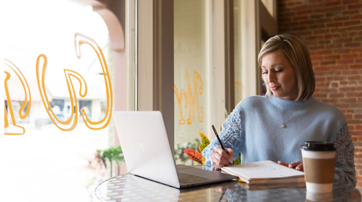 Photo of female employee writing in notebook and working on her laptop while sitting inside of a cafe.