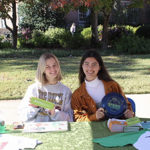 Two smiling student interns sitting at a green table holding a reusable utensil kit and a frisbee.