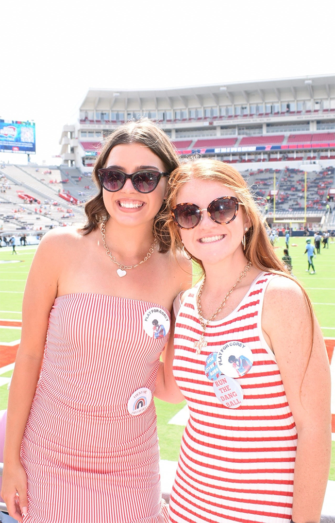 Two young women stand on the sidelines in a football stadium on a sunny day.