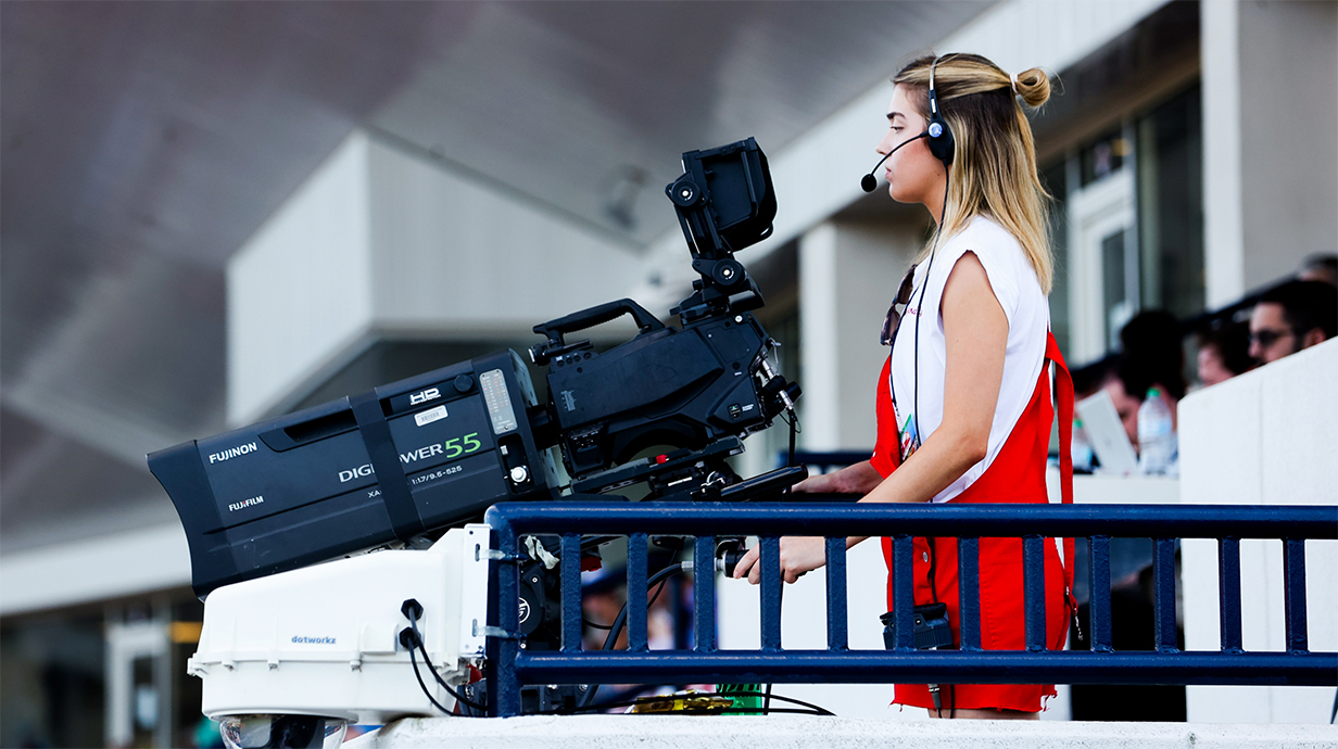 A young woman wearing red overalls operates a TV camera in a stadium.