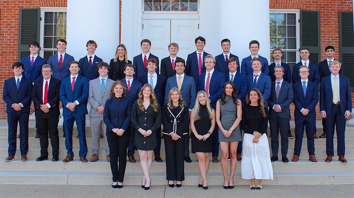 A large group of young people stands on the steps of a massive brick building with white columns.