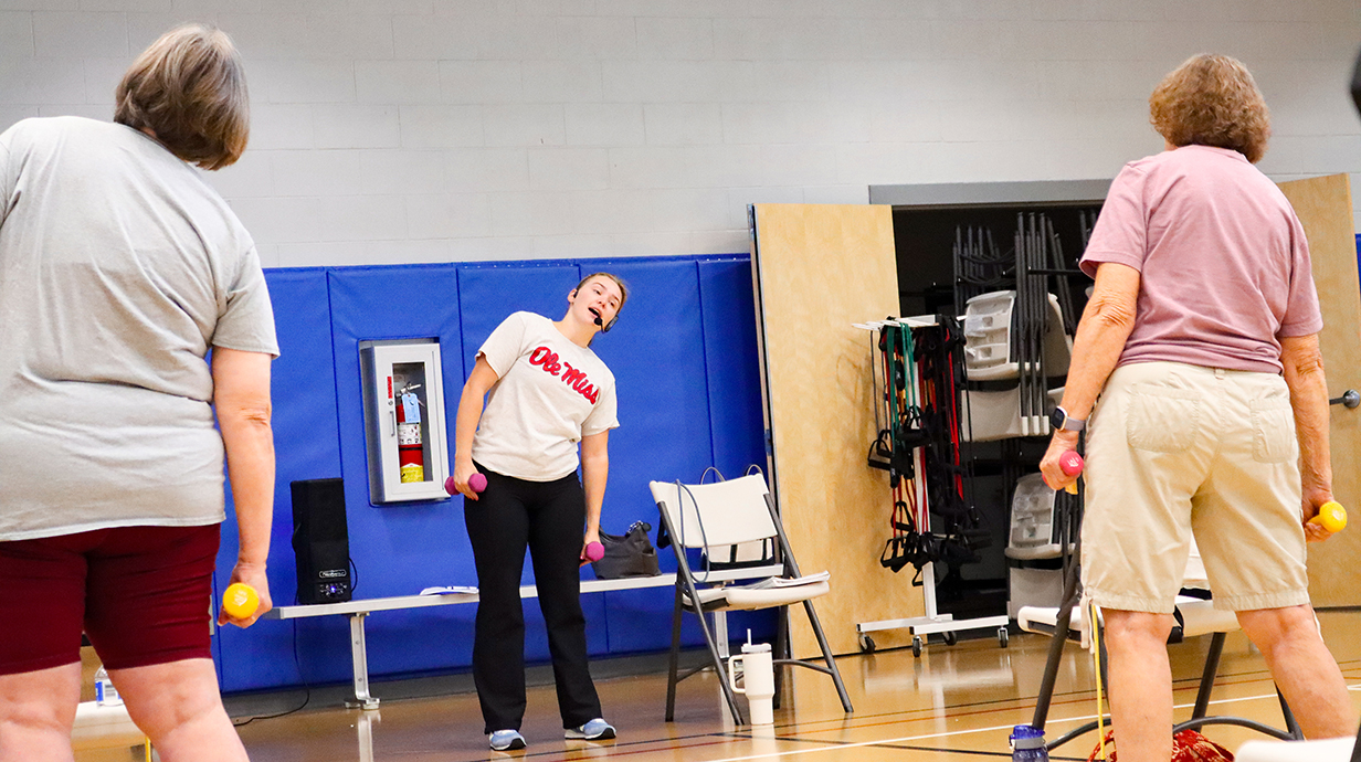 A young woman leads an exercise class for a group of older women in a gym.