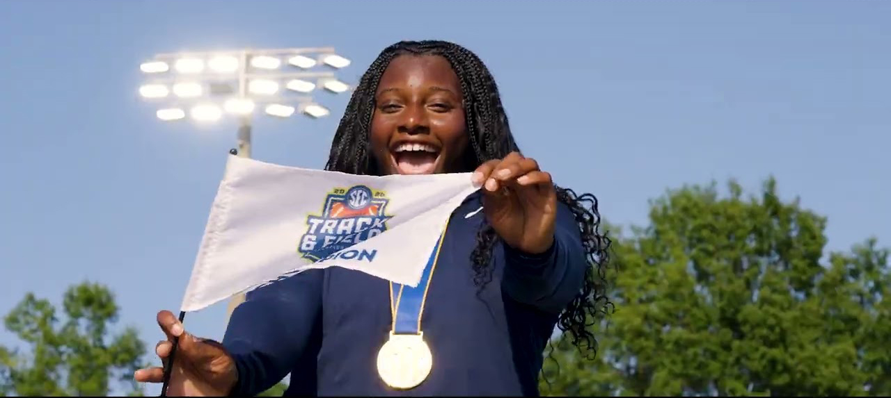A smiling track and field athlete holds up a championship flag and wears a gold medal after winning an SEC track and field championship.