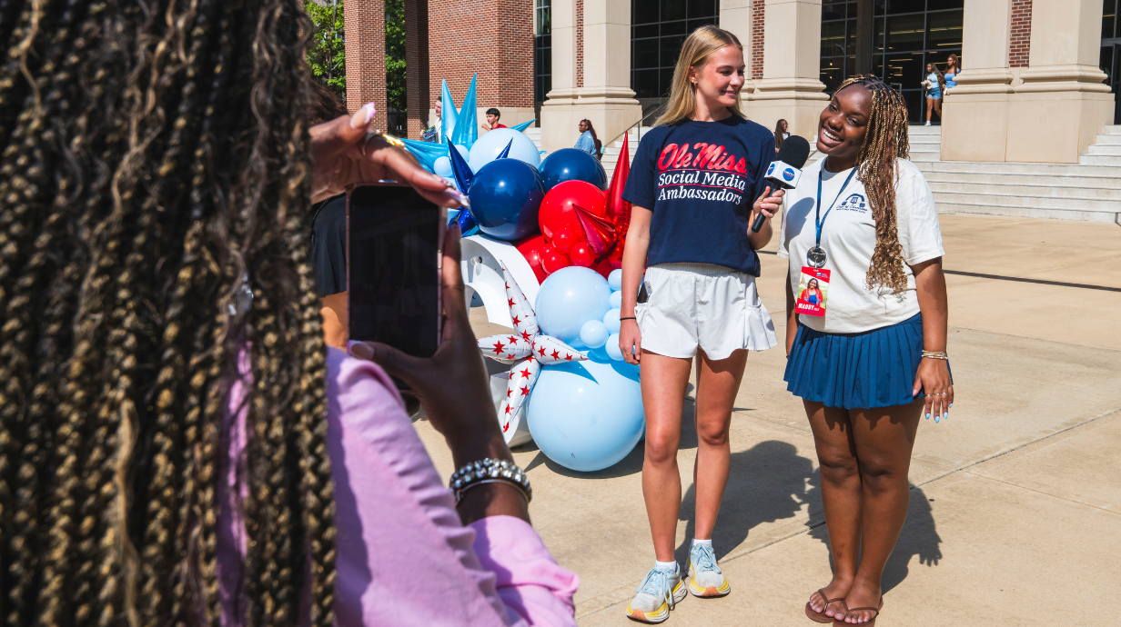 A student wearing an Ole Miss Social Media Ambassadors shirt holds a microphone while interviewing another student in front of the Union.