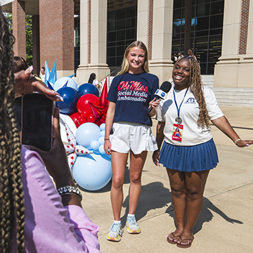 Social Media Ambassador holding a microphone with another student outside a campus building.
