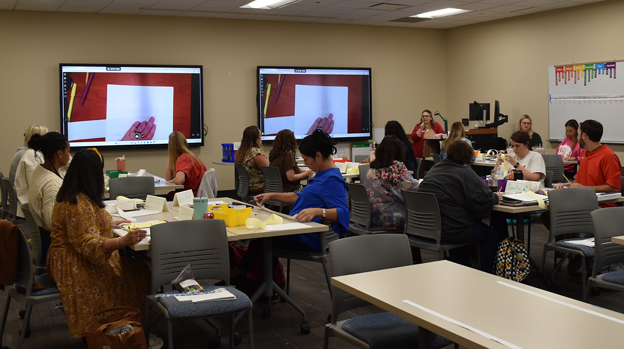 A group of educators in a classroom setting, attentively watching a demonstration on large screens.