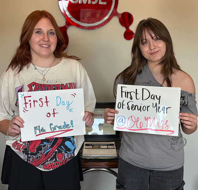 Two female students stand side by side smiling at the camera while holding mini white board signs.