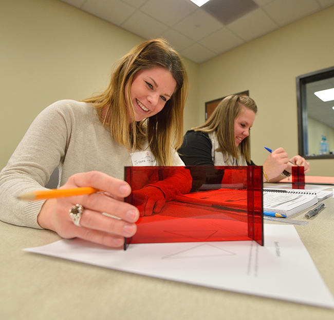 Two female teachers sit at a table engaged in a fun activity using red transparent shapes and paper