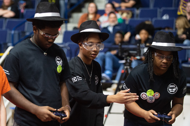 Three male students using remote controllers to move a robot (not seen).