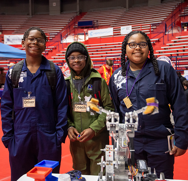 Three students in uniforms smile proudly beside a robotics project in a gymnasium setting.