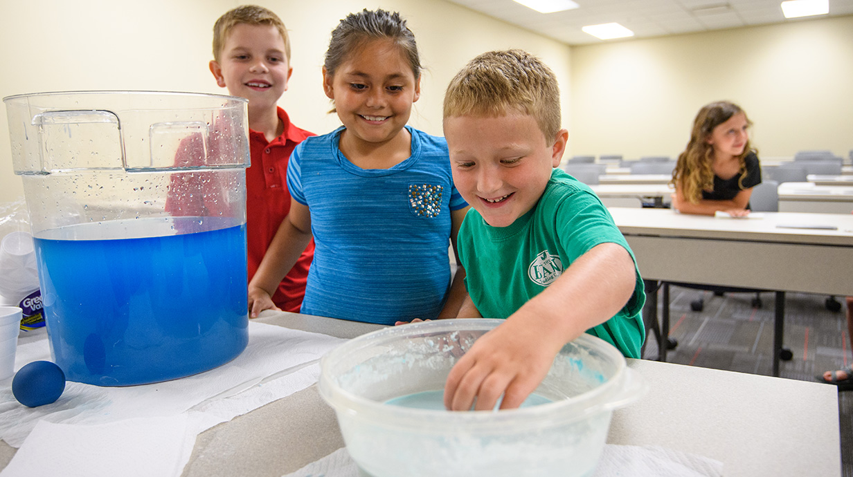 Three children are in a classroom, smiling and engaged in a science activity with one boy reaching into a bowl of blue liquid.