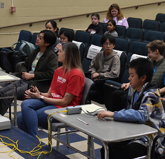 two students sit desks for a math competition with a moderator in the middle