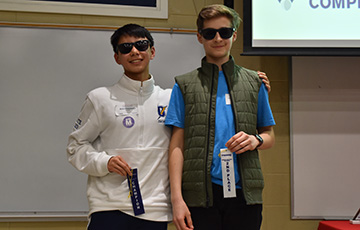 Two young men wearing sunglasses stand side by side, smiling, and holding award ribbons with a "Math Competition" banner partially visible behind them.