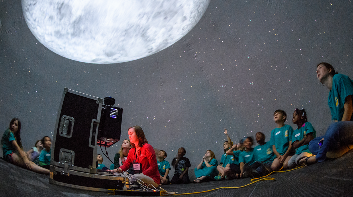 A group of children sits in a planetarium, gazing at a large projection of the moon above.