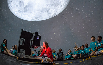 A group of children sit inside a planetarium dome, gazing at a large moon projection with a woman operating the console.