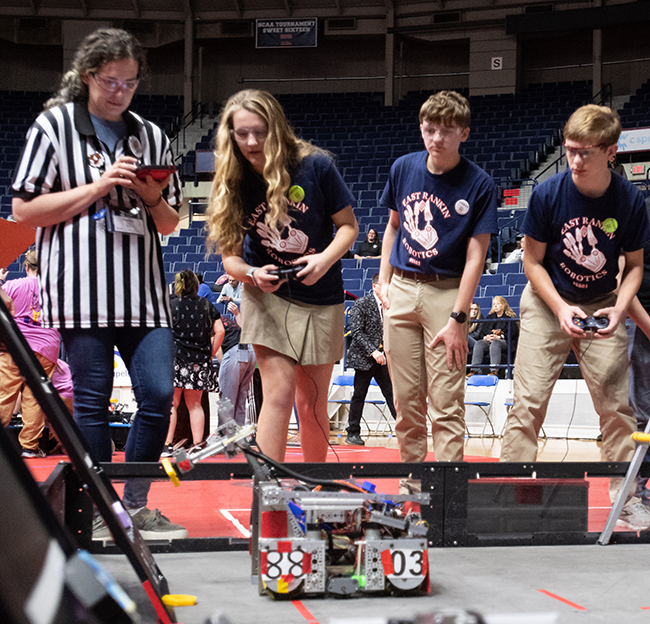 Students operate a small competition robot on a game field while a referee records scores, inside a large arena with spectators in the background.