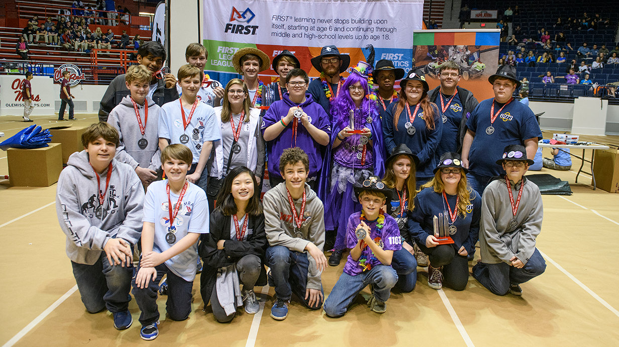 A group of students and mentors, wearing medals and colorful outfits, proudly pose at a robotics competition with a FIRST Robotics banner behind them.