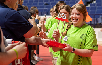 A smiling participant in a bright green event shirt is handed a small red trophy, with others in matching shirts and a crowd in an arena behind them.