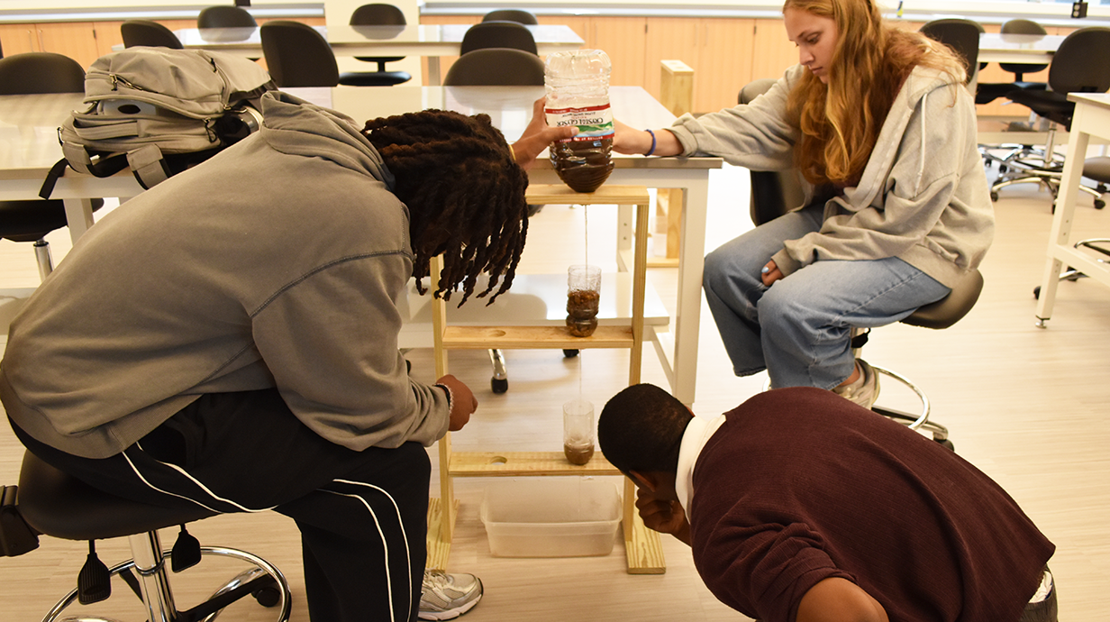 Three students conduct a science experiment in a classroom, using a water filtration setup with plastic bottles.