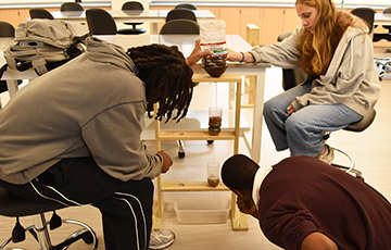 Three students conduct a science experiment in a classroom, using a water filtration setup with plastic bottles.