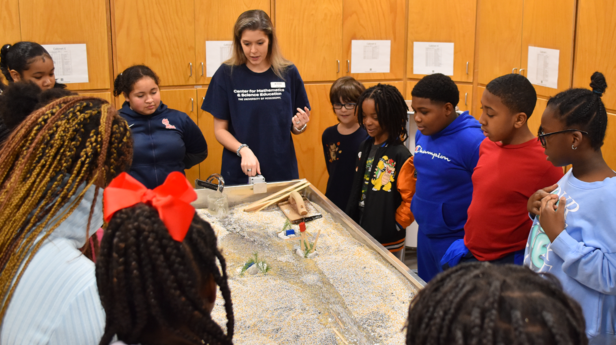 A teacher stands with a group of children around a stream table model, demonstrating a water flow experiment.