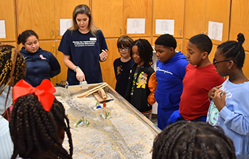 A teacher stands with a group of children around a stream table model, demonstrating a water flow experiment.
