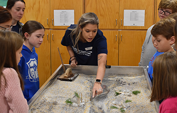 Female graduate students points out the water's pathway on the stream table while students gather around and watch.