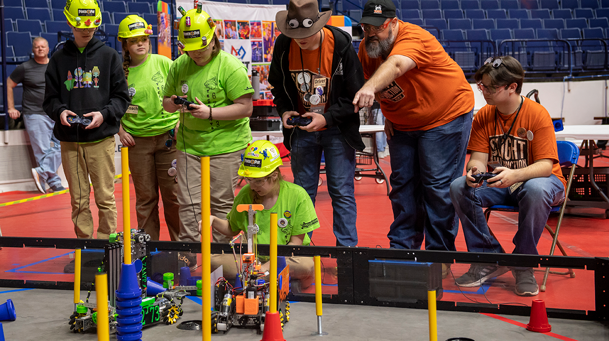 A group of students and mentors operate and observe robots on a competition field, with several participants in bright green shirts and hard hats controlling robots while others watch and point, inside a large arena.