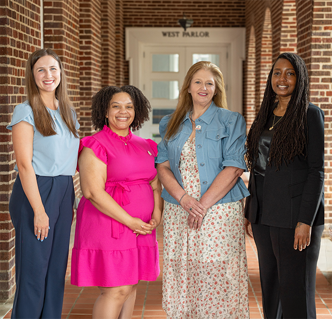 Four women stand smiling in a hallway with brick walls.