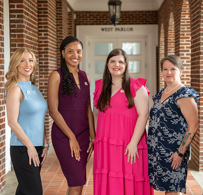 Four women stand smiling in a hallway with brick walls.