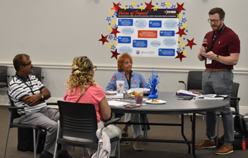 A man presents to a group seated at a round table in a conference room. A colorful poster with stars is on the wall, creating an engaging atmosphere.