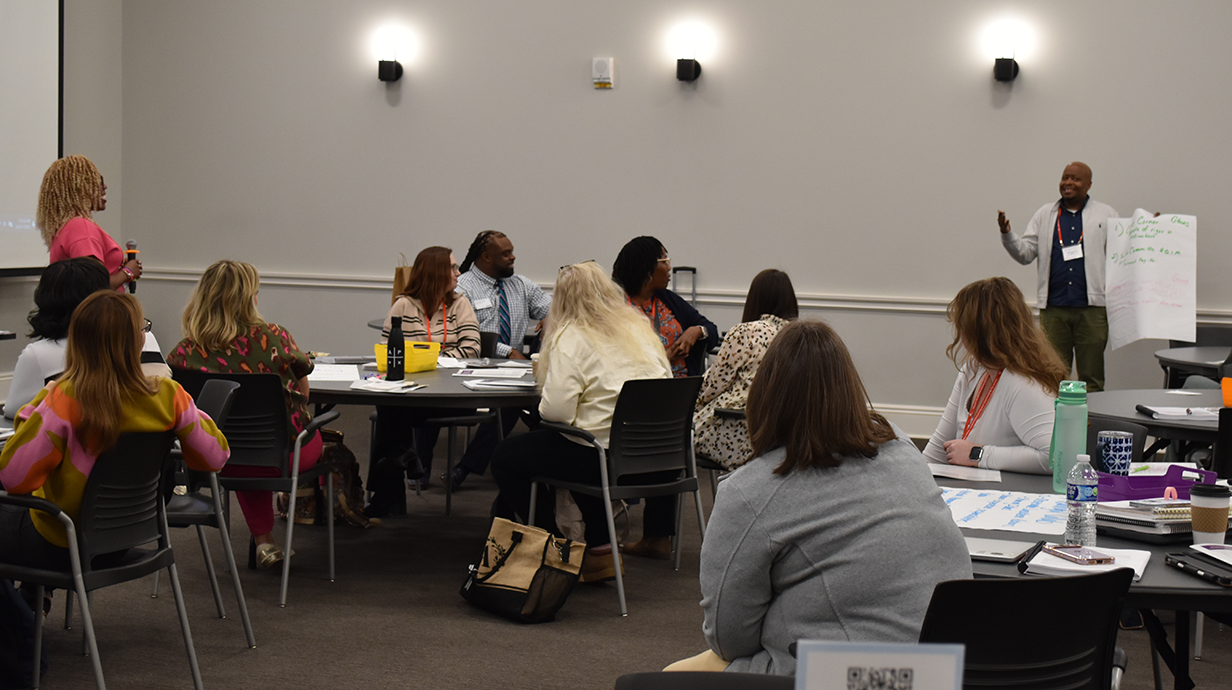 A group of math teachers sit at tables in a conference room, listening to a speaker presenting with a large notepad.