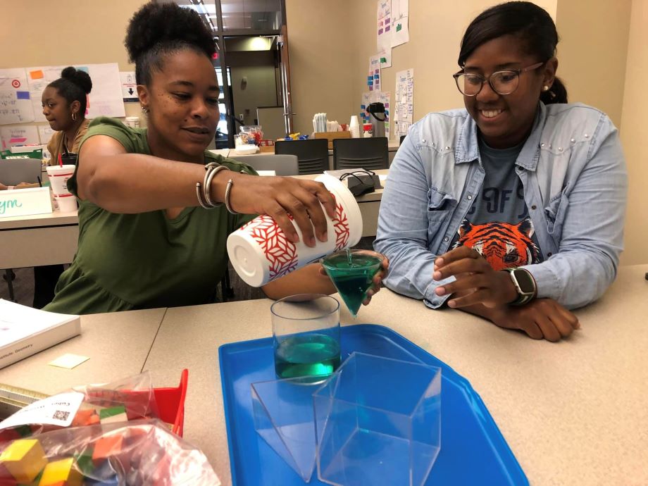 Two women in a classroom are engaged in an activity that explore volume, pouring a green liquid into a triangular container.