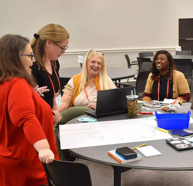 A group of women discussing mathematics learning around a table filled with papers and notebooks at the Annual Mathematics Specialist Conference.