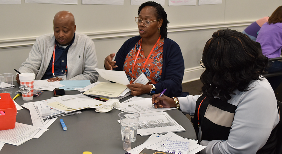 Three math educators engaged in a discussion around a cluttered table with papers and pens, conveying a collaborative and focused atmosphere.