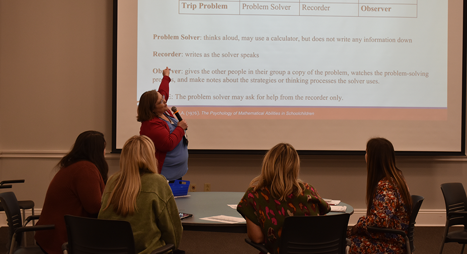 A woman in a red sweater points at a presentation slide with a microphone while four seated women face her