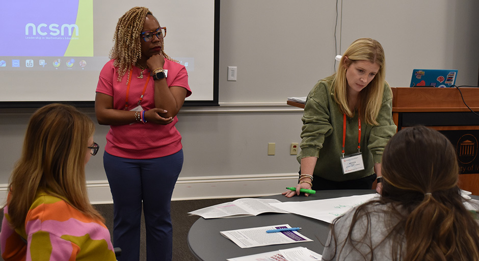 Two women lead a discussion in a conference room. One stands with arms crossed, listening intently, while the other leans over a table covered with papers.