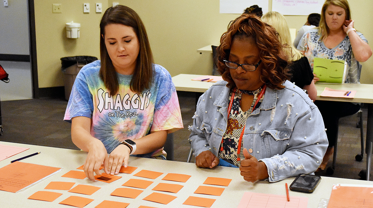 Two women sit at a table engaged in a card-sorting activity. The woman on the left wears a tie-dye shirt, while the woman on the right wears a denim jacket. 