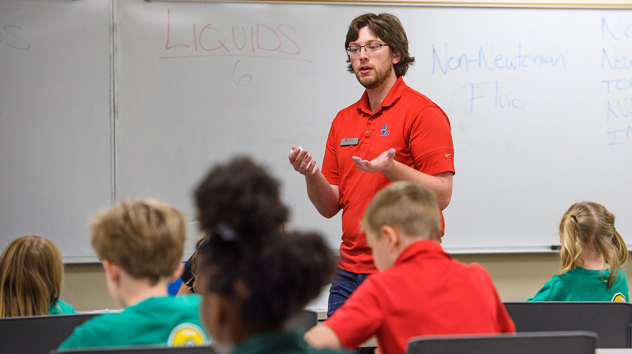 A man in a red shirt stands in front of a whiteboard speaking to a classroom of young students seated at desks, focused and engaged.