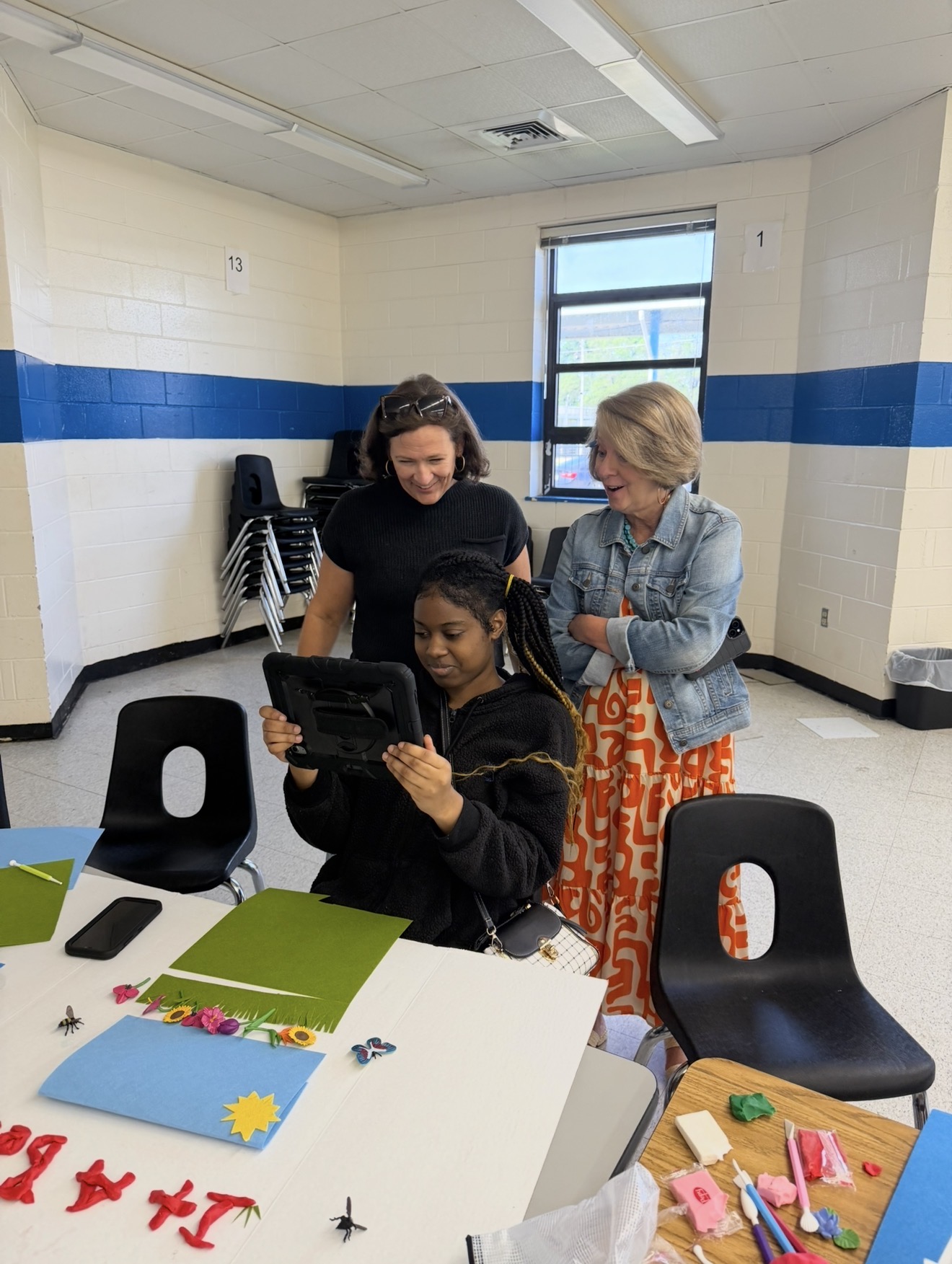 A student uses a tablet to showcase a completed classroom project while two teachers stand nearby, smiling and observing. The table is covered with colorful craft materials, paper, and modeling clay used to create the project.